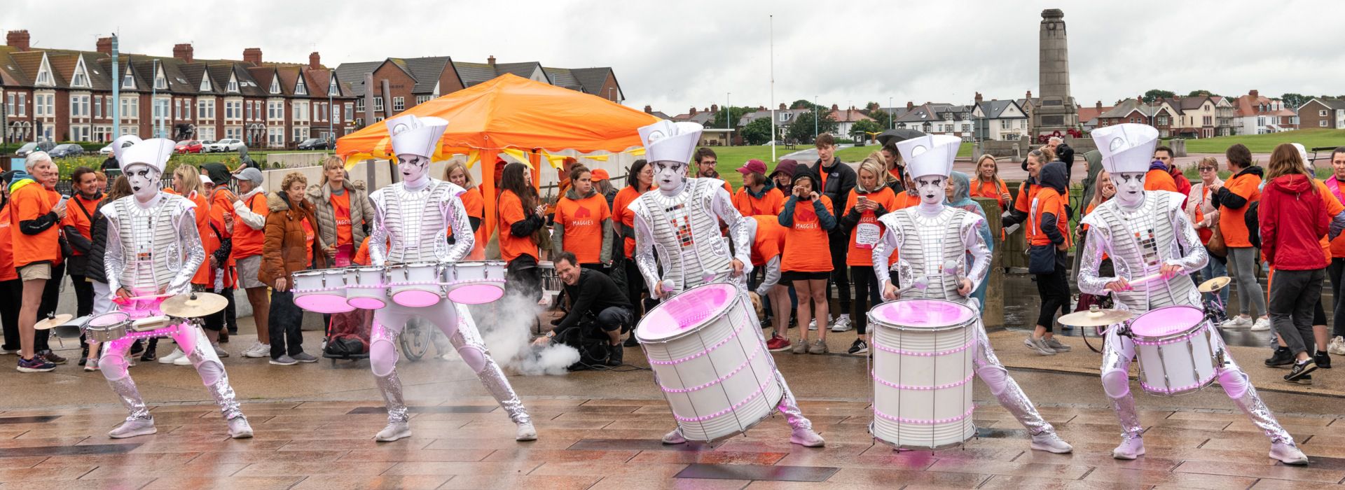 Drummers in costumes performing at coastal crawl with Maggie's supporters in background wearing orange