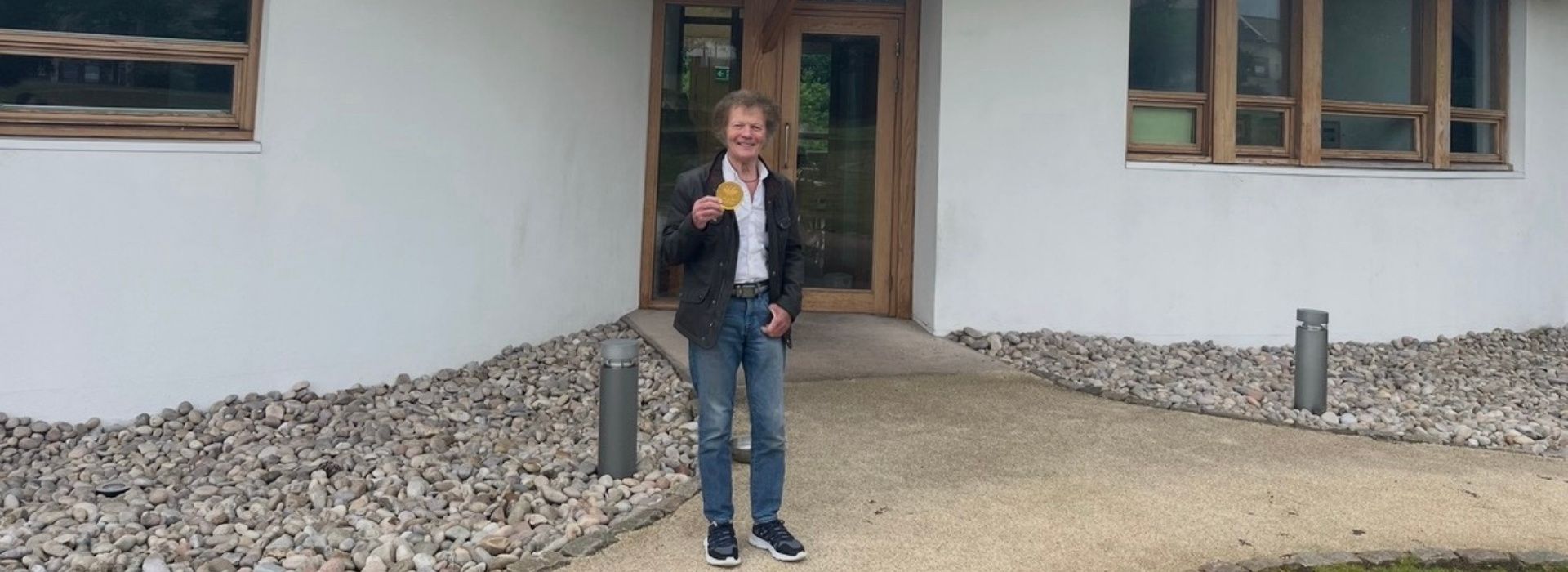 Man (Peter) stands holding medal smiling in front of Maggie's Dundee cancer centre