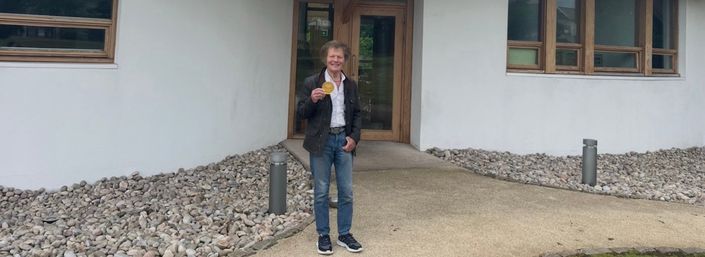Man (Peter) stands holding medal smiling in front of Maggie's Dundee cancer centre